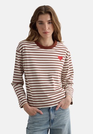 Jonge vrouw met een wit shirt met lange mouwen en bruine horizontale strepen, met een klein rood hartje op de borst, gecombineerd met lichtblauwe spijkerbroek.
