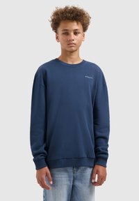 Tienager met krullend haar, gekleed in een marineblauwe sweatshirt met kleine witte tekst en lichtblauwe jeans, staand tegen een effen achtergrond.