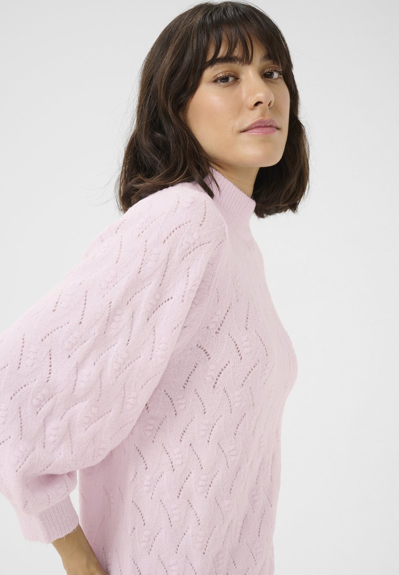 Maglione lavorato a maglia rosa chiaro con collo alto e motivo testurizzato. Presenta maniche leggermente a sbuffo e polsini a coste allungati.