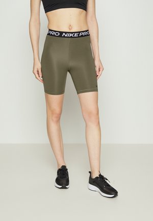Donna che indossa pantaloncini da ciclismo Nike Pro color oliva, reggiseno sportivo nero e scarpe da corsa nere su uno sfondo semplice.