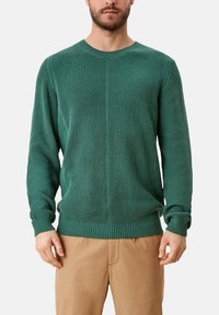 Homme portant un pull vert à côtes en maille à manches longues et un pantalon marron clair, debout devant un fond blanc uni.