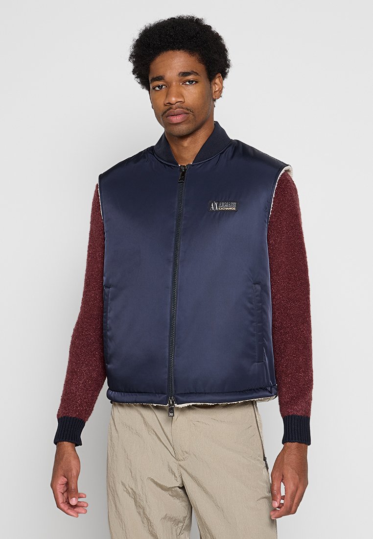Giovane uomo con capelli afro che indossa un giubbotto senza maniche blu navy sopra un maglione bordeaux e pantaloni beige, in piedi davanti a uno sfondo neutro.