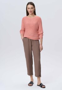 Jasnoróżowy sweter dzianinowy z szerokimi rękawami, zestawiony z luźnymi brązowymi spodniami. Modelka nosi czarne płaskie sandały i minimalną biżuterię.