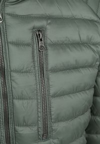 Donkergroene pufferjack met een gewatteerde textuur, voorzien van een ritszak op de linkerborst en zwarte ritshardware.