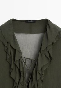 Olivenfarvet bluse med flæsekrave, lavet af letvægtsstof. Den har en V-udskæring og en snørefunktion foran.