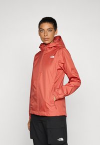Water-resistant, hooded jacket in coral color with zip front, elastic cuffs, and logo detail on chest; smooth, lightweight fabric.
