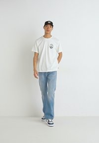 Hvit kortermet t-skjorte med rund hals og et logo på brystet, kombinert med lyseblå straight-leg jeans og svarte sneakers.