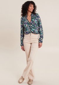Langärmlige Bluse mit einem lebhaften Paisleymuster in Blau, Grün und Weiß, kombiniert mit hellbeigen Hosen, die über Frontknöpfe und Seitentaschen verfügen.