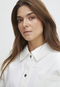 Hvid knap-op jakke lavet af tekstureret stof, med en spids krave, sorte knapper og en brystlomme detalje.