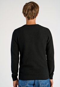 Schwarzer gestrickter Pullover mit strukturiertem Muster, Rundhalsausschnitt und langen Ärmeln; kombiniert mit blauen Jeans, was einen lässigen Look ergibt.