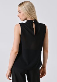 Donna bionda che indossa una blusa nera senza maniche con apertura a chiave sul retro e bottone, abbinata a pantaloni neri, che guarda a sinistra su uno sfondo semplice.
