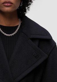 Černý vlněný kabát s širokými klopami, podrobným prošíváním a logem. V kombinaci s černým pleteným topem a masivním stříbrným náhrdelníkem.