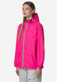 Donna in piedi che indossa una giacca con cappuccio rosa acceso con cerniera frontale e pantaloni cargo grigio chiaro, rivolta leggermente verso destra.