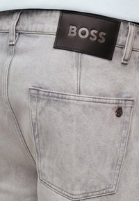 Πίσω τσέπη από ανοιχτόγκρι τζιν με μικρό μεταλλικό λογότυπο και μαύρο δερμάτινο ταμπελάκι με την επωνυμία "BOSS" στη ζώνη μέσης.