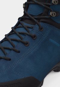 Μπλε μπεζές πεζοπορικό μποτάκι με μαύρα μεταλλικά οπές και μαύρα κορδόνια που περνούν μέσα από γάντζους και κρίκους.