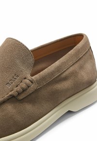 Brauner Wildleder-Slipper mit weißer Gummisohle, detailreicher Naht und geprägtem "BOSS"-Logo auf der Oberseite nahe der Zehenpartie.