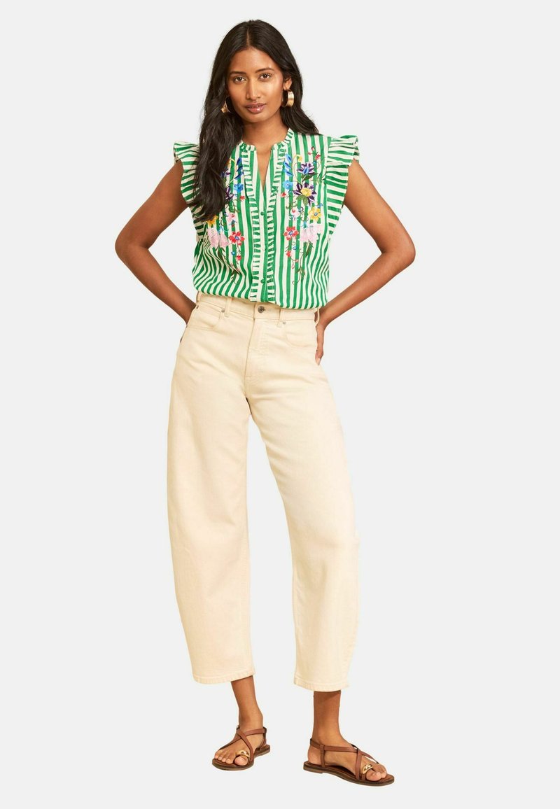 Woman standing with hands on hips, wearing a green striped embroidered top, wide-leg cream pants, and brown sandals, against a plain background.