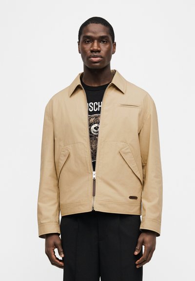 Άνδρας που φοράει μπεζ ζακέτα με φερμουάρ πάνω από μαύρο πουκάμισο με σχέδιο αρκούδας, συνδυασμένο με μαύρο παντελόνι, στέκεται μπροστά σε ένα απλό φωτεινό φόντο.