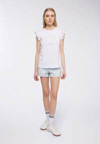 Top bianco a maniche corte con maniche a balze, caratterizzato da un design con taglio a cuore e decorazioni. Abbinato a pantaloncini di jeans e scarpe bianche.