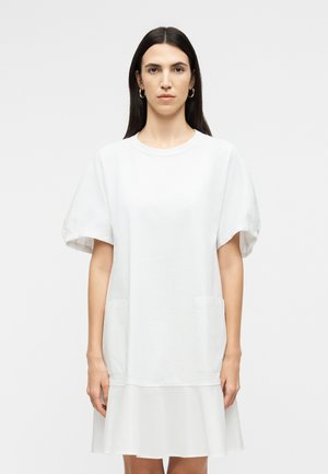 Γυναίκα με μακριά σκούρα μαλλιά, ντυμένη με λευκό φόρεμα κοντών μανικιών με τσέπες και βολάν στο τελείωμα, στέκεται μπροστά σε απλό ανοιχτόχρωμο φόντο.