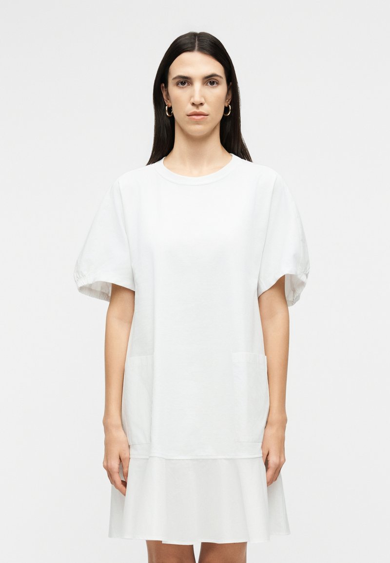 Γυναίκα με μακριά σκούρα μαλλιά, ντυμένη με λευκό φόρεμα κοντών μανικιών με τσέπες και βολάν στο τελείωμα, στέκεται μπροστά σε απλό ανοιχτόχρωμο φόντο.