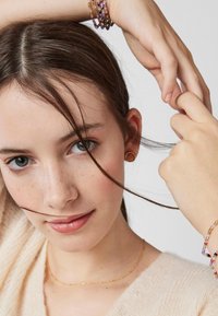 Złote kolczyki w kształcie kół, delikatny naszyjnik z złotej łańcuszka i kolorowe bransoletki z koralików na modelce z brązowymi włosami i jasną cerą.