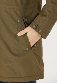 Olivgrön jacka med en texturerad tyg, tryckknappsbrytningar och en sidofick, med en åtsittande design vid handleden.