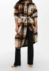 Longue surchemise à carreaux dans des tons crème, marron et vert. Dispose d'une fermeture à boutons sur le devant, de deux poches poitrine et d'une ceinture à nouer à la taille.