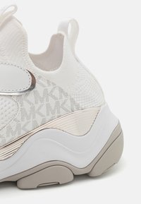 Scarpa da ginnastica bianca con tomaia in maglia testurizzata, dettagli metallici e suola in gomma solida con design ondulato e dettagli grigio chiaro.
