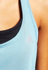 Lyseblå atletisk tanktop med rund halsudskæring, lagdelt over en sort tanktop. Har et subtilt adidas logo i lyserød på fronten.