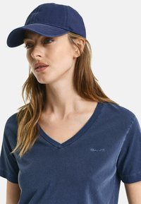 Marineblå bomulds-kasket med en buet skygge og syede detaljer, båret med en matchende marineblå v-hals t-shirt med et lille logo på brystet.