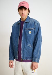 Denim jaka ar klasisku griezumu, gaiši zila auduma, priekšējā rāvējslēdzēja aizdare, neliela krūšu kabata un izšūta Carhartt logo kreisajā pusē.