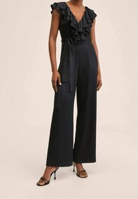 Femme portant une combinaison noire sans manches avec un décolleté en V à volants et des jambes larges, associée à des sandales à talons noires.