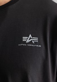 Fekete pamut póló, ezüst logóval, három vízszintes vonallal fent, valamint stilizált "A" és "I" betűkkel, és az "ALPHA INDUSTRIES" szöveggel alatta.
