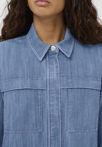 Svijetloplava traper jakna s klasičnom kragnom, dugmadi na zatvaranju, dva džepa na prsima i suptilnim detaljima šivanja. Mekana tekstura.