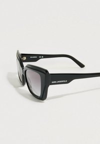 Černé hranaté sluneční brýle s gradientními čočkami, tlustými plastovými obroučkami a jemným logem na nožičce. Hladká textura a výrazný tvar.