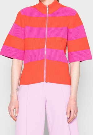 Kvinde iført en ribbet strikket orange og lyserød stribet zip-up bluse med ærmer til albuerne og lyserøde bukser, stående indendørs.