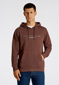 Brun huvtröja med framficka, dragsko i huva och tryckt "COPENHAGEN LINDBERGH"-logotyp. Mjuk tyg och avslappnad passform.