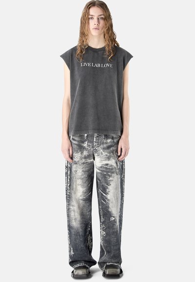 Jeune personne aux cheveux longs portant un haut sans manches gris foncé avec le texte "LIVE LAB LOVE" et un pantalon large noir et blanc délavé et usé.
