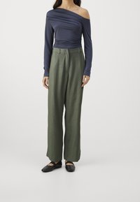 Haut à manches longues bleu marine avec détails froncés, associé à un pantalon large taille basse vert olive. Chaussures plates noires complètent la tenue.