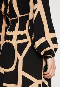 Schwarz-beiges Kleid mit geometrischem Muster, elastischem Bund und Puffärmeln. Der Stoff wirkt leicht und hat eine glatte Textur.