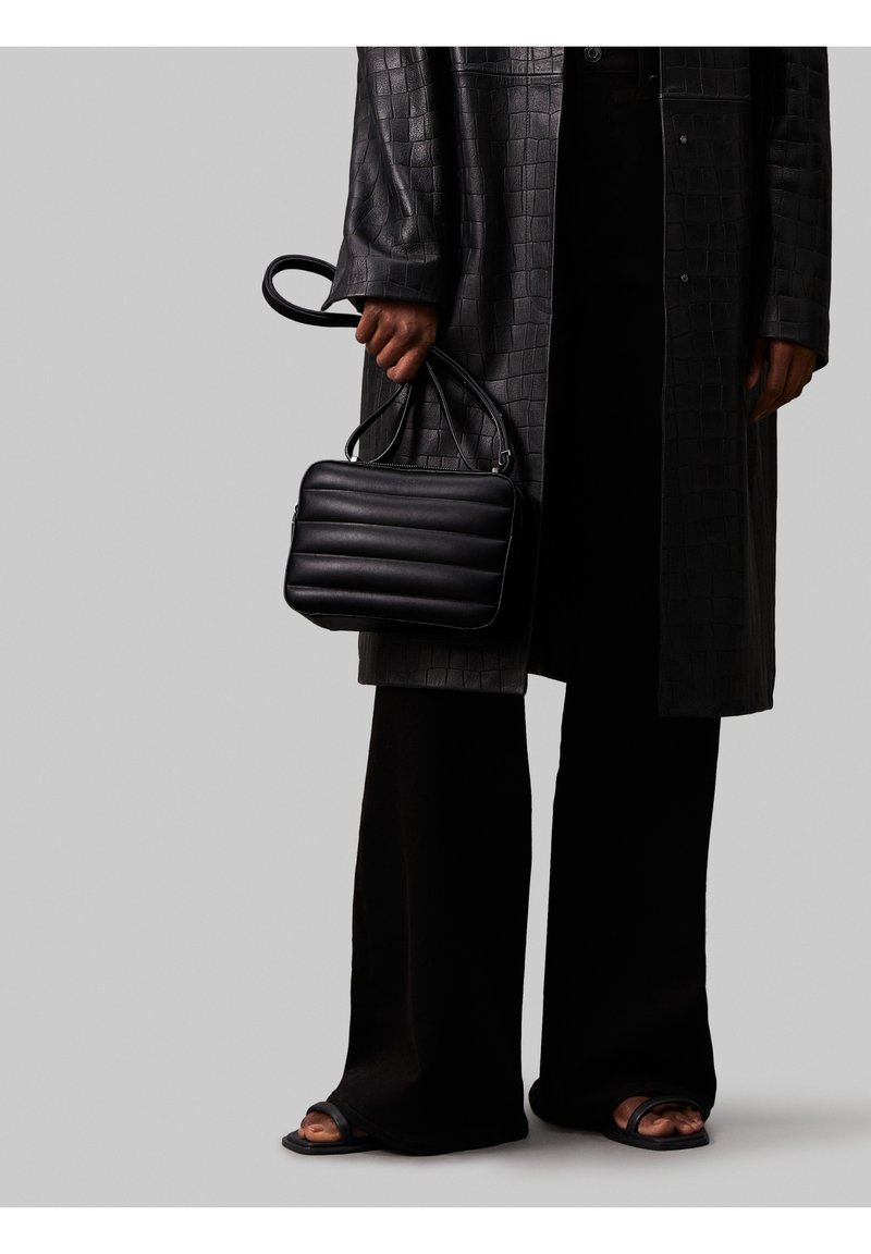 Czarna skórzana torebka z pikowaną teksturą, prostokątny kształt, podwójne uchwyty. Nosi się ją z długim czarnym, teksturowanym płaszczem i szerokimi czarnymi spodniami.