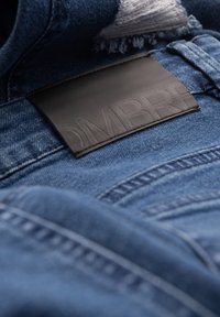 Primer plano de unos pantalones de mezclilla azul con una etiqueta de marca de cuero negro cosida en la cintura, mostrando la textura detallada de la tela y las costuras.