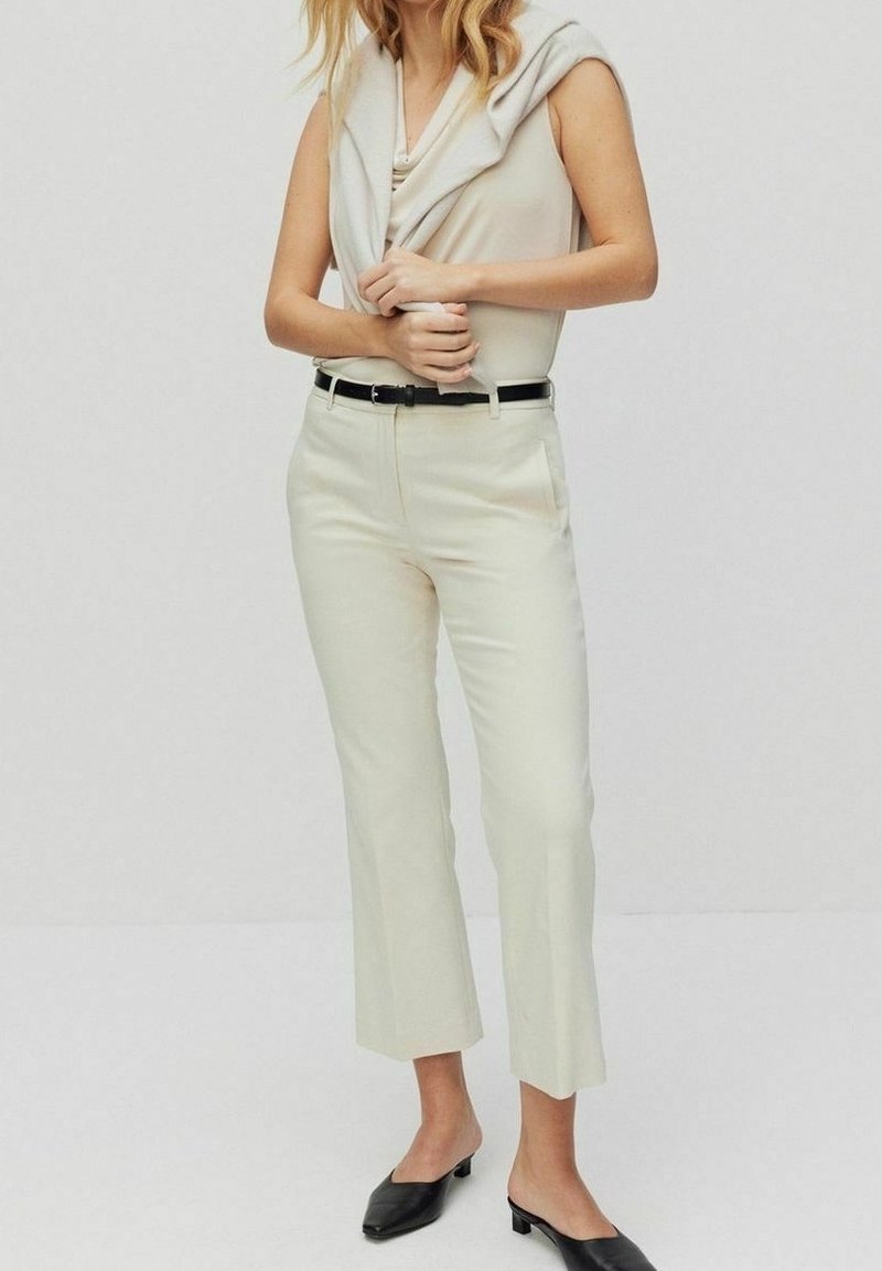 Femme portant un haut beige sans manches avec un pantalon beige raccourci assorti, une ceinture noire et des mules noires à petit talon, debout devant un fond uni.