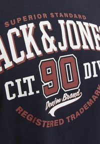 Tamnoma modra bombažna majica z belim in burgundskim bold napisom. Oblika vključuje "SUPERIOR STANDARD," "90" in "DENIM BRAND" v različnih stilih.