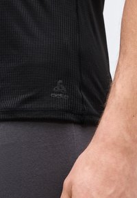 Gros plan sur le torse et le bras d'une personne portant une chemise noire côtelée avec un petit logo près de l'ourlet et un pantalon gris foncé.