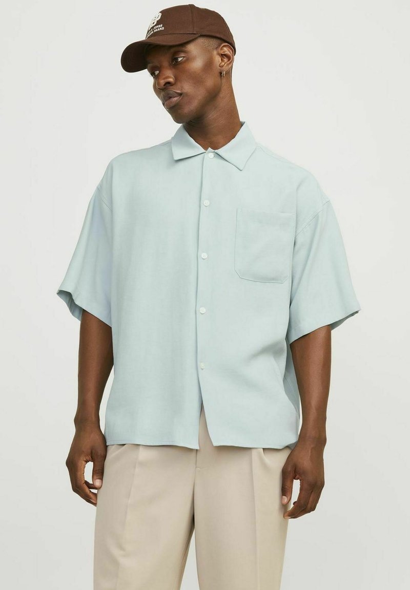 Camisa de manga corta azul claro con cuello, que presenta un bolsillo en el pecho y una textura sutil. Llevada con pantalones beiges y una gorra marrón.