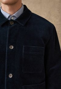 Giacca di velluto a coste blu navy con chiusura a bottoni, colletto a punta e taschino sul petto. Presenta una texture a coste e bottoni lisci.