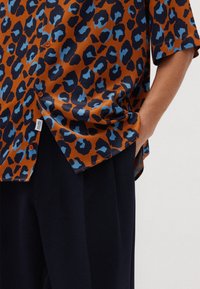 Camicia in tessuto arancione con stampa leopardata caratterizzata da macchie blu scuro e blu chiaro. Maniche corte, vestibilità ampia e patta dei bottoni.