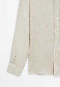 Chemise beige à manches longues avec poignet boutonné et patte de boutonnage à l'avant, montrée de la moitié de la manche jusqu'à l'ourlet inférieur sur un fond uni.